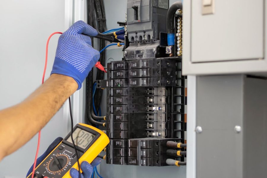 Electrician wearing blue gloves using a digital multimeter to test wiring inside a circuit breaker panel