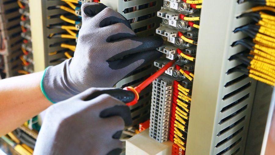 Person wearing protective gray gloves tightens a red wire onto a terminal strip inside an electrical control panel with yellow cables in the background.