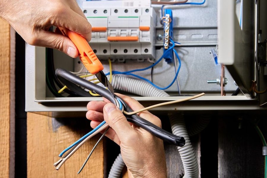 Close-up of a person stripping and connecting electrical wires inside a breaker panel using a bright orange tool.